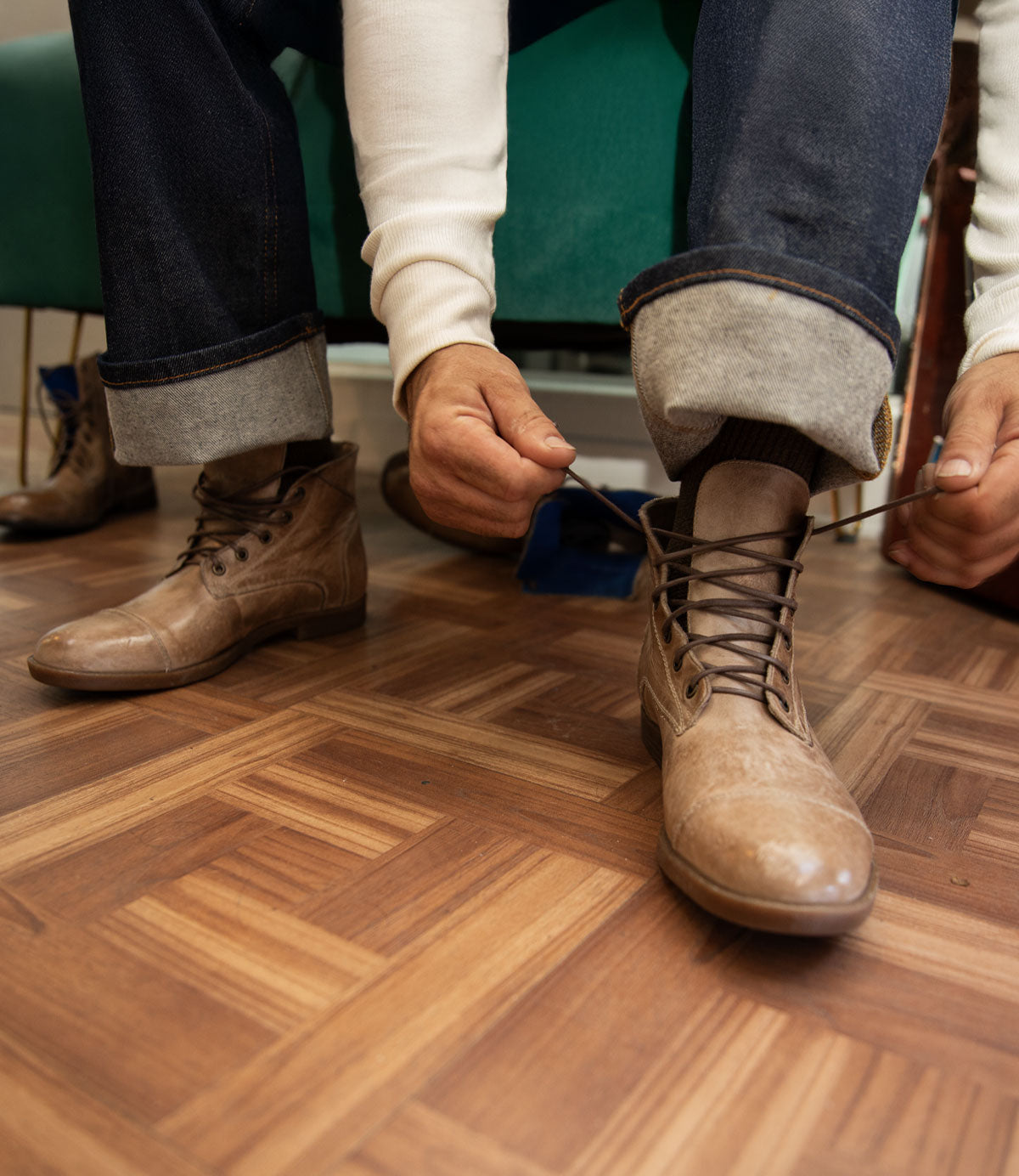 A person in rolled-up jeans and a white shirt is sitting and tying the laces of a brown leather Leonardo boot by Bed|Stü, which features a flexible rubber outsole, on a wooden floor.