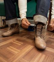 A person in rolled-up jeans and a white shirt is sitting and tying the laces of a brown leather Leonardo boot by Bed|Stü, which features a flexible rubber outsole, on a wooden floor.