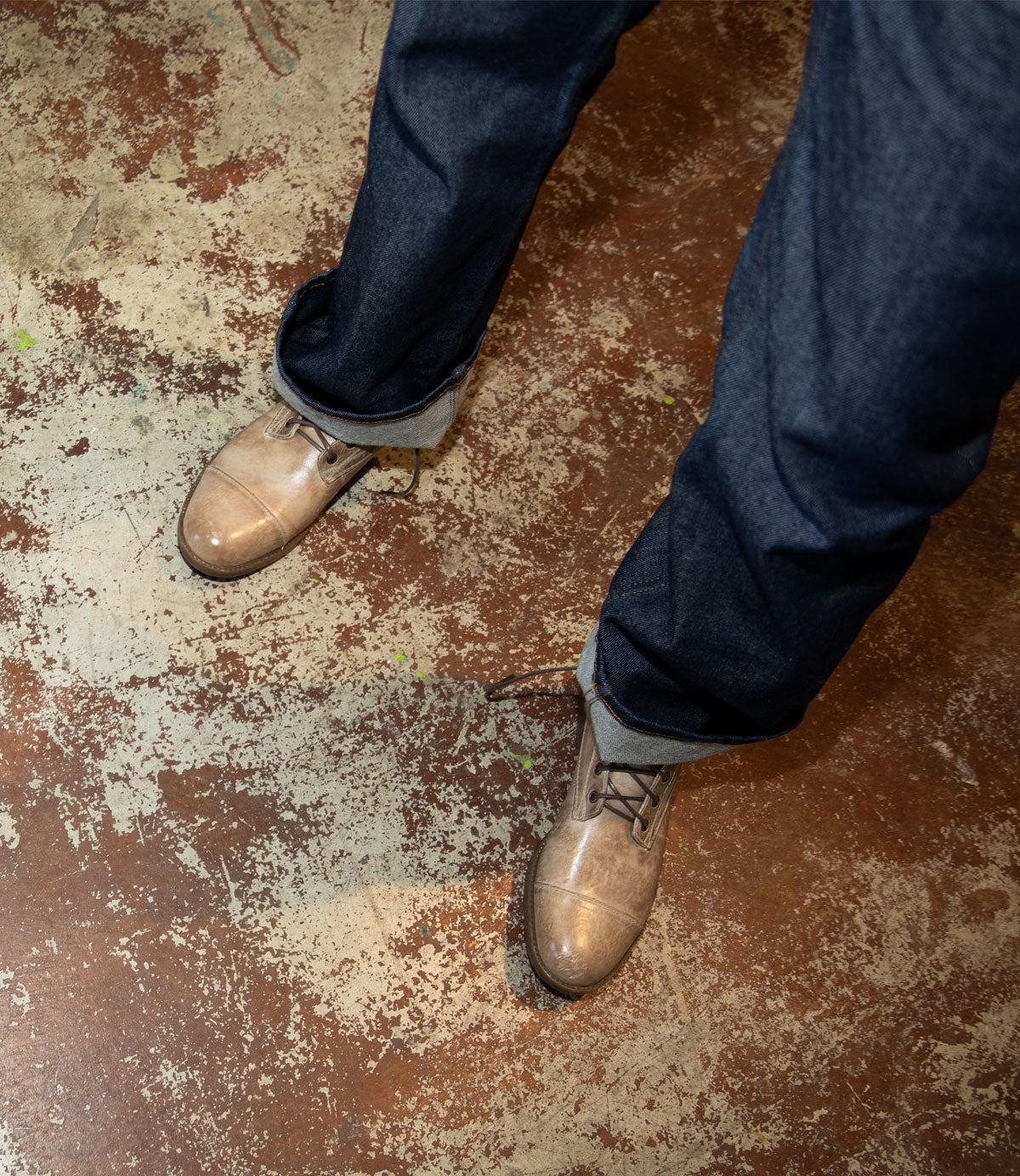 A person wearing cuffed dark blue jeans and Bed|Stü’s Leonardo light brown leather boots with a cushioned insole stands on a worn, textured brown floor.