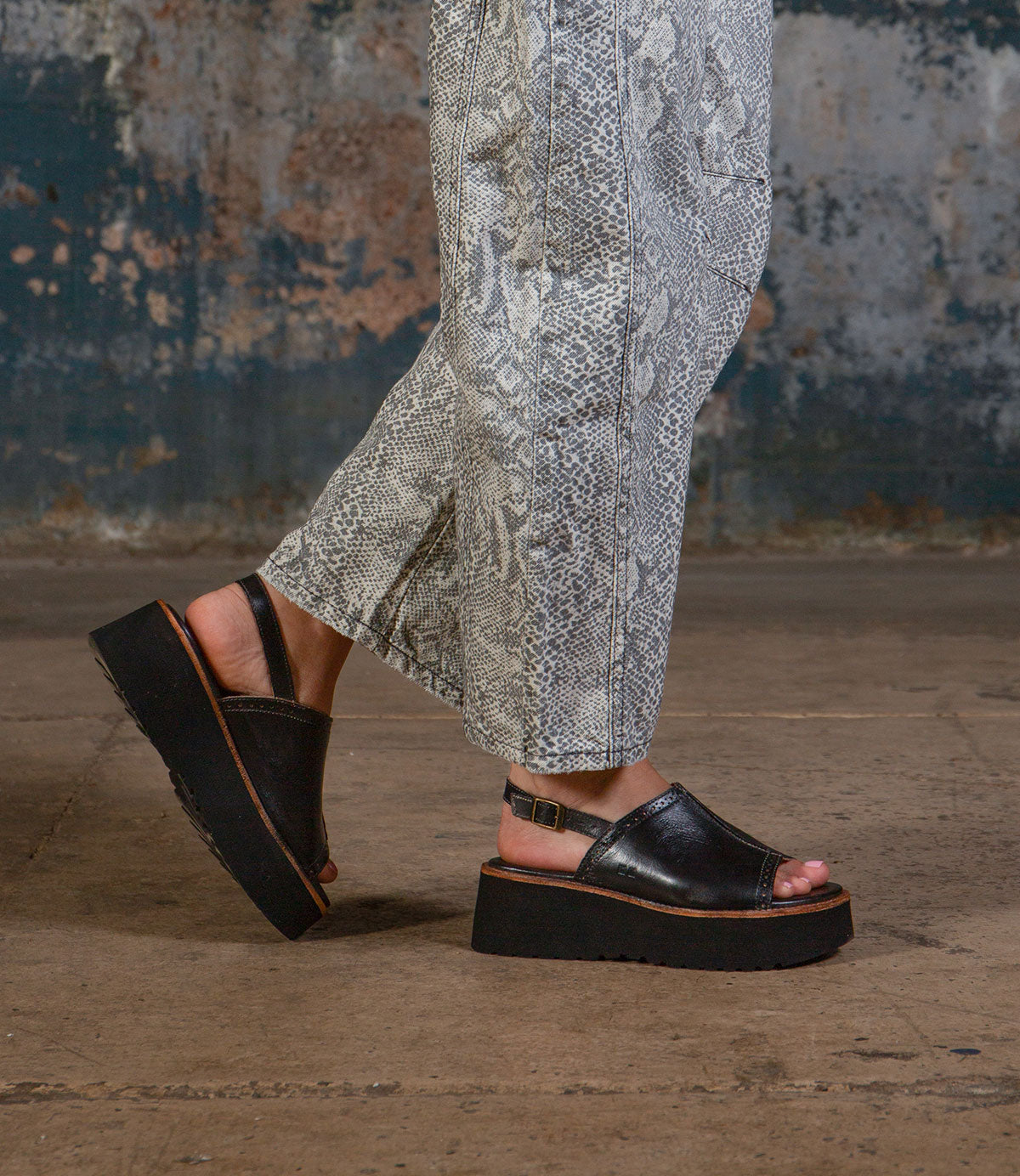 A person wearing the Bed|Stü Library black platform sandals—a perfect nod to retro footwear and ‘90s style—paired with snake print pants, stands on a concrete floor against a worn, textured wall.