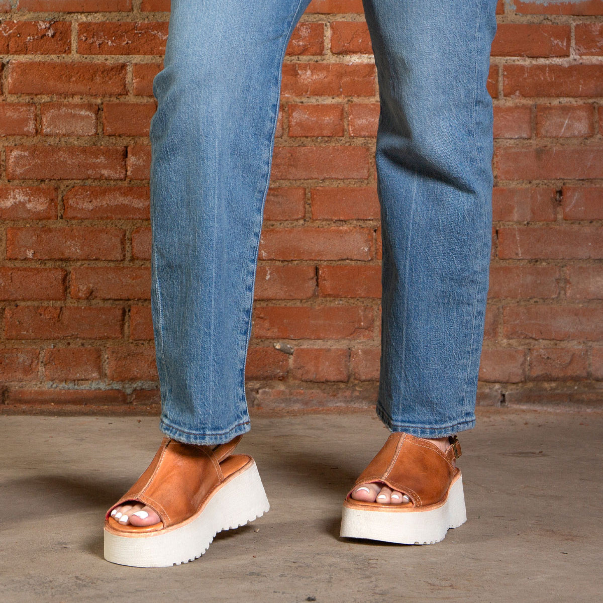 Person wearing blue jeans and Bed|Stü Library brown platform sandals with white soles, standing on a concrete floor in front of a brick wall—a perfect nod to ‘90s style with this retro footwear choice.