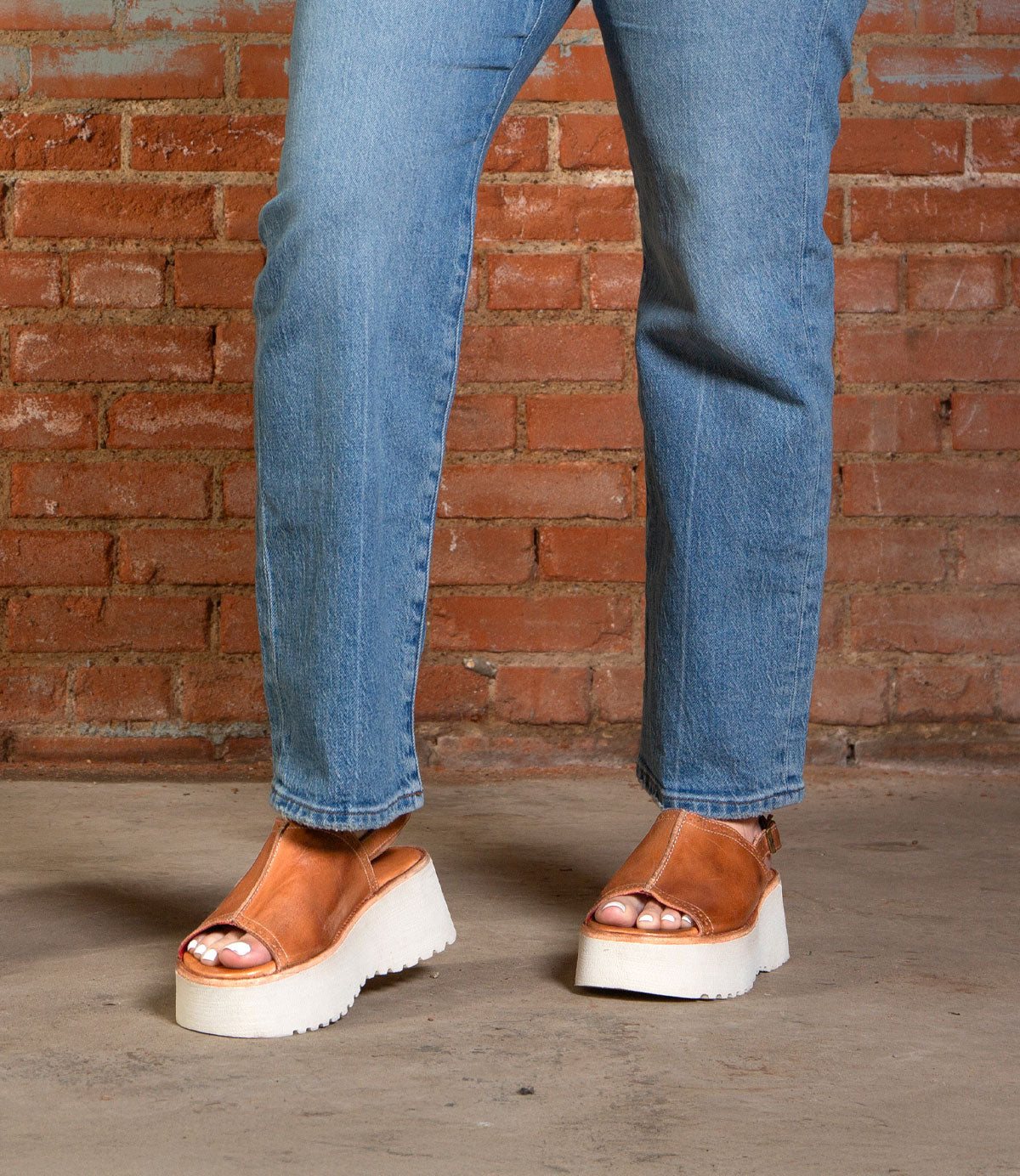 Person wearing blue jeans and Bed|Stü Library brown platform sandals with white soles, standing on a concrete floor in front of a brick wall—a perfect nod to ‘90s style with this retro footwear choice.