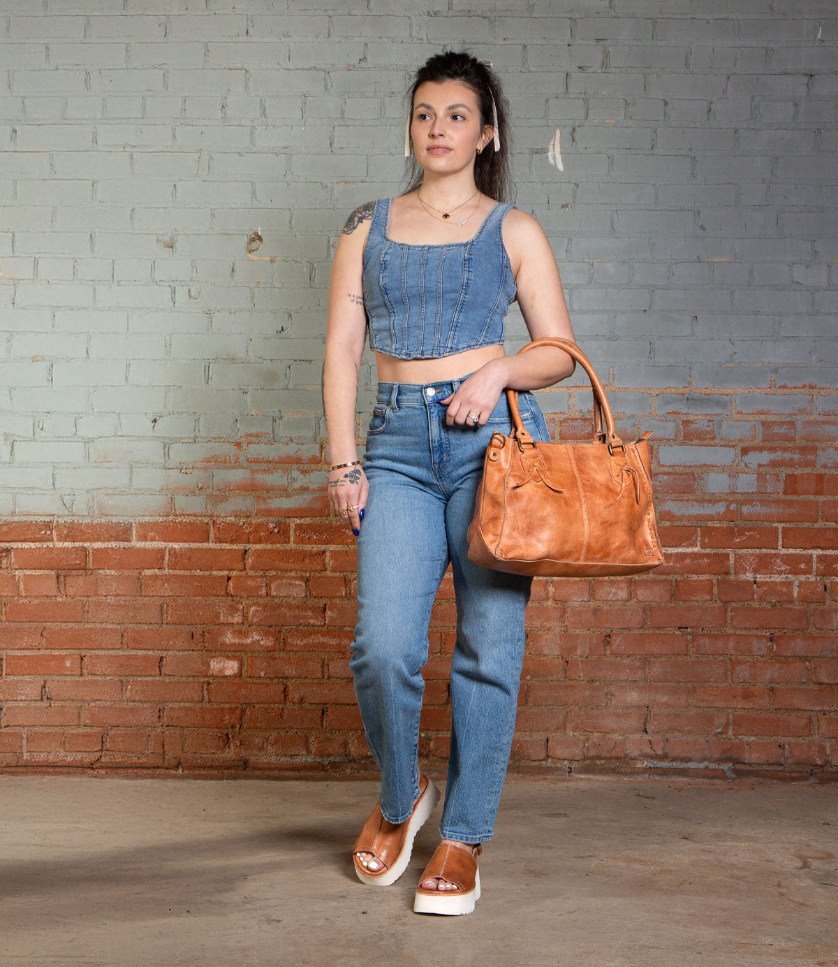 A woman carrying the Bed|Stü Library large tan handbag, styled with a sleeveless denim crop top, high-waisted jeans, and tan retro platform sandals, stands against a brick and painted wall.