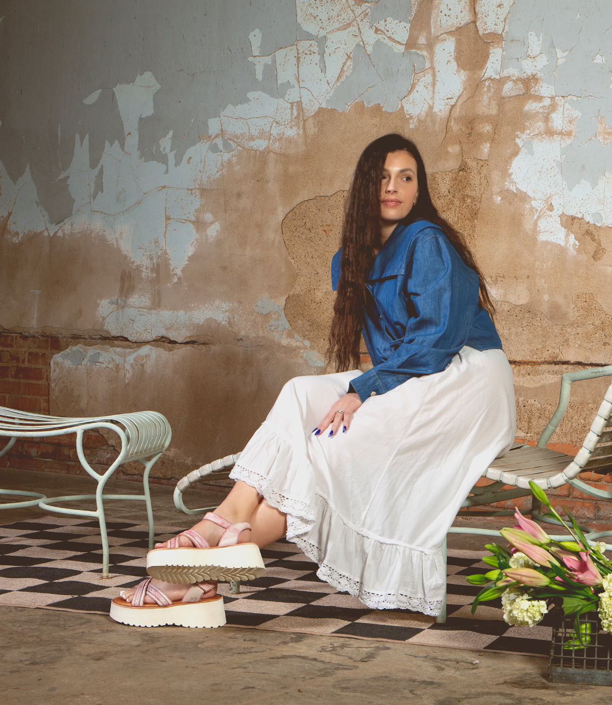 A woman in a denim jacket and white skirt sits on a metal bench indoors, surrounded by peeling walls and a tiled floor, with a bouquet of flowers nearby—her feet elegantly crossed in comfortable Bed|Stü Lift sandals.