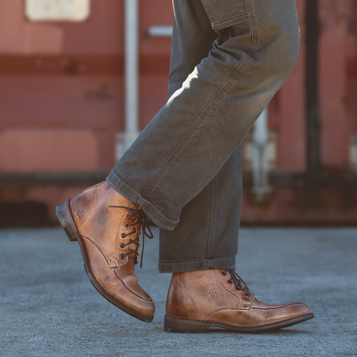 A person wears the Bed|Stü Lincoln II brown leather lace-up men's boots with an apron toe and dark gray pants, standing on a concrete surface with a reddish-brown background.