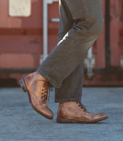 A person wears the Bed|Stü Lincoln II brown leather lace-up men's boots with an apron toe and dark gray pants, standing on a concrete surface with a reddish-brown background.