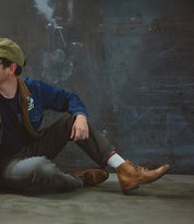 A person in a denim jacket, olive pants, and Bed|Stü Lincoln II men's boots with an apron toe sits on the ground against a dark, textured wall.