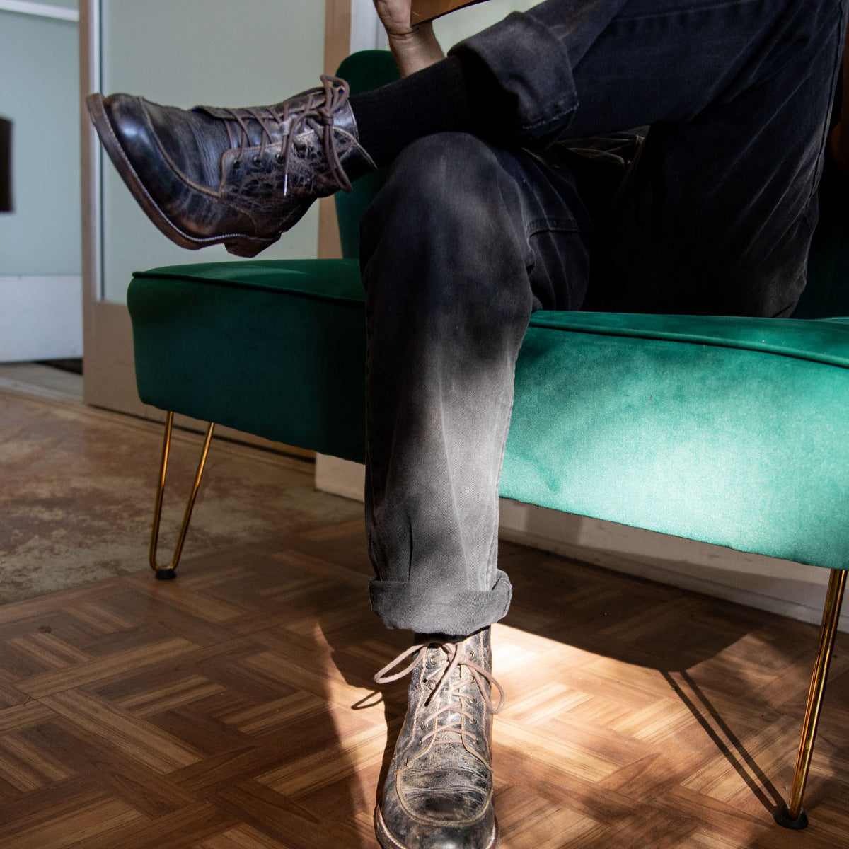 A person sits on a green sofa with their legs crossed, holding a closed book and wearing black jeans paired with Bed|Stü Lincoln II men's work-boot style lace-up boots featuring an apron toe. Sunlight illuminates the parquet floor and part of the Bed|Stü boot.