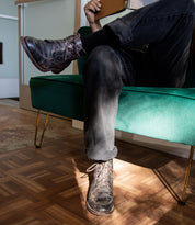 A person sits on a green sofa with their legs crossed, holding a closed book and wearing black jeans paired with Bed|Stü Lincoln II men's work-boot style lace-up boots featuring an apron toe. Sunlight illuminates the parquet floor and part of the Bed|Stü boot.