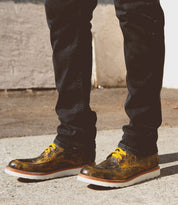 Person wearing black pants and metallic-colored Bed Stu Lincoln boots with yellow laces, standing outdoors on a concrete surface.