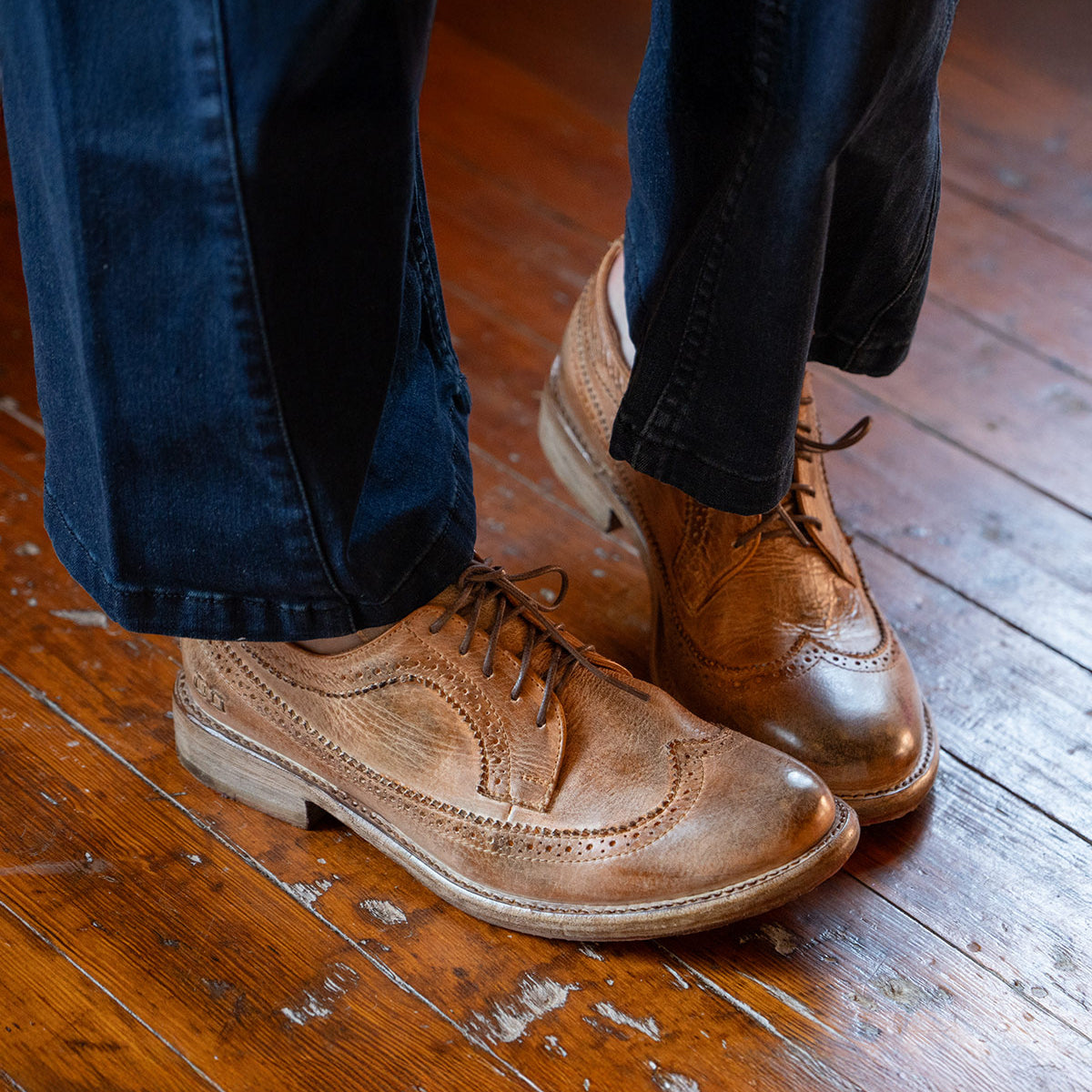 Wearing the Bed|Stü Lita K II classic derby shoes featuring brogue detailing and a wingtip toe, paired with dark blue pants, standing on a worn wooden floor.
