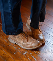 Wearing the Bed|Stü Lita K II classic derby shoes featuring brogue detailing and a wingtip toe, paired with dark blue pants, standing on a worn wooden floor.