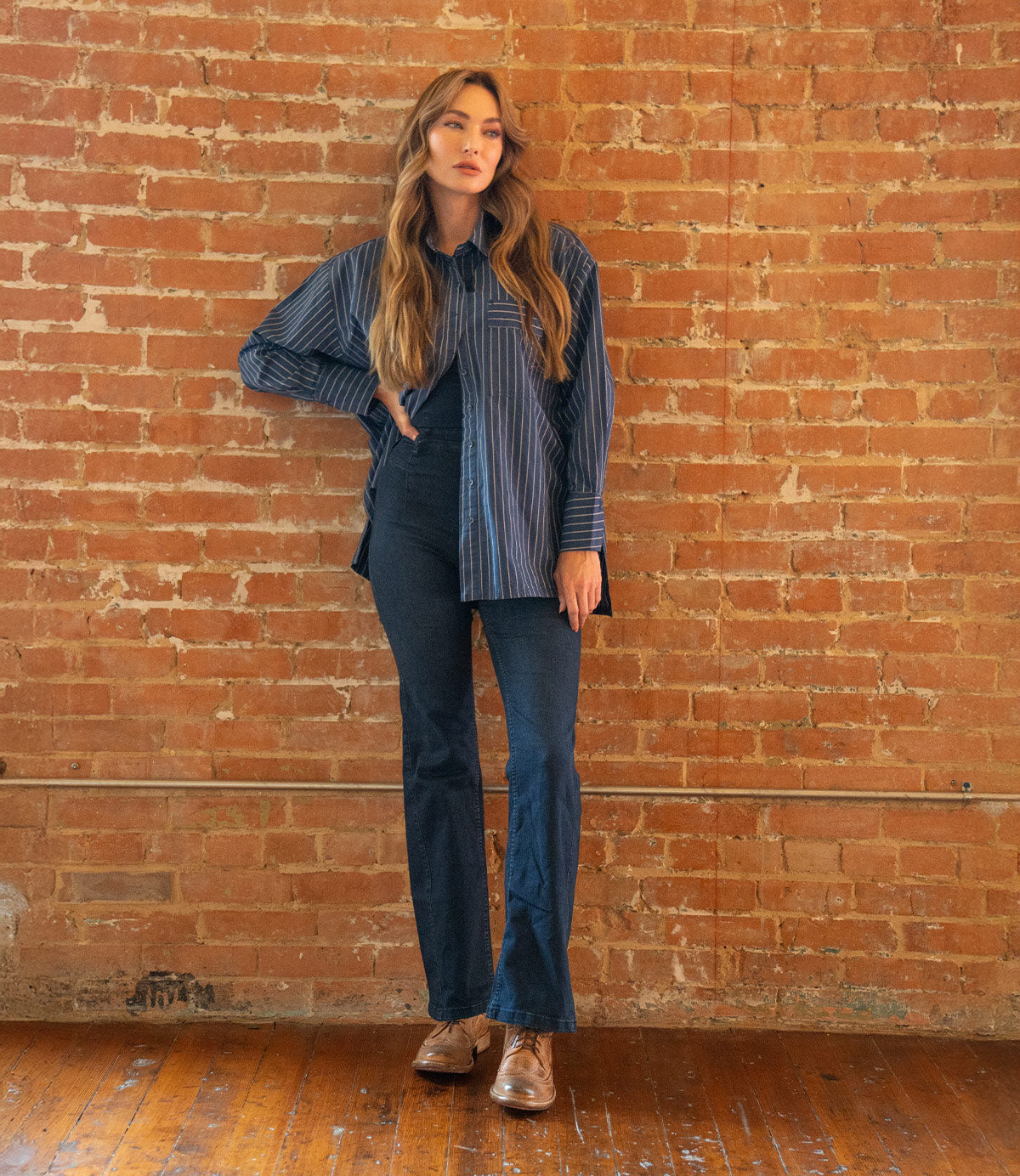 A person stands indoors against a brick wall, dressed in a blue striped shirt and dark blue pants, wearing the Bed|Stü Lita K II brown shoes featuring classic derby styling and subtle brogue detailing, with one hand on their hip.