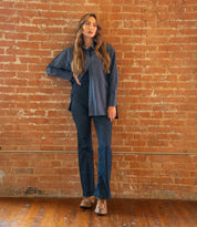 A person stands indoors against a brick wall, dressed in a blue striped shirt and dark blue pants, wearing the Bed|Stü Lita K II brown shoes featuring classic derby styling and subtle brogue detailing, with one hand on their hip.