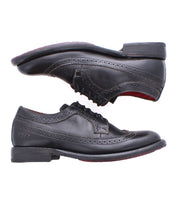 Two black leather Lita K II dress shoes by Bed|Stü, featuring brogue detailing and a wingtip toe, are displayed from the side; one classic derby is upright while the other rests above it with its sole visible.