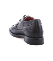 The Bed|Stü Lita K II is a black leather classic derby dress shoe with refined brogue detailing and a wingtip toe, shown from the back left angle against a white background.