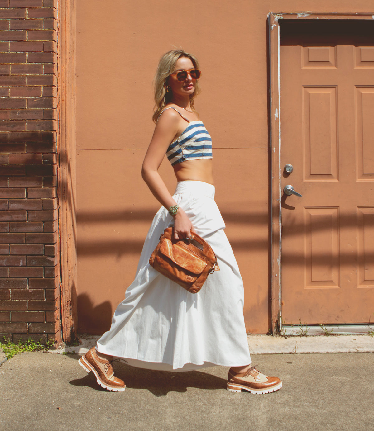 A woman in sunglasses, a striped crop top, white skirt, and tan shoes walks past a tan door holding a brown bag; her look is finished with the classic touch of Bed|Stü’s Lita K III vintage-inspired shoes.
