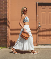 A woman in sunglasses, a striped crop top, white skirt, and tan shoes walks past a tan door holding a brown bag; her look is finished with the classic touch of Bed|Stü’s Lita K III vintage-inspired shoes.