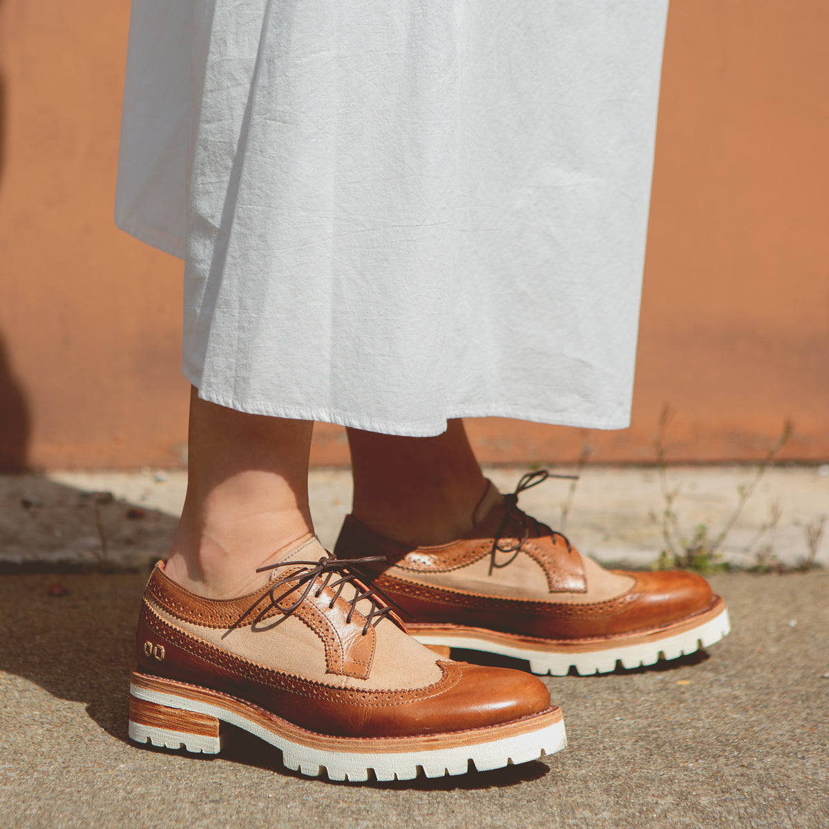A person stands on a sidewalk wearing a long white skirt and Bed|Stü Lita K III brown canvas and leather brogue shoes with thick white soles.