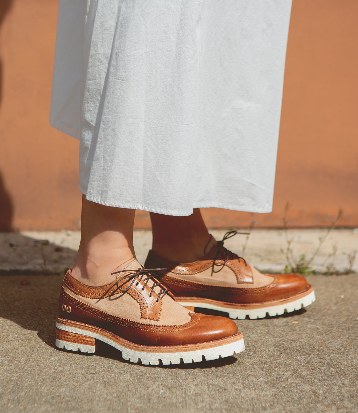 A person stands on a sidewalk wearing a long white skirt and Bed|Stü Lita K III brown canvas and leather brogue shoes with thick white soles.