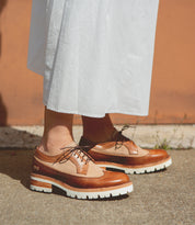 A person stands on a sidewalk wearing a long white skirt and Bed|Stü Lita K III brown canvas and leather brogue shoes with thick white soles.