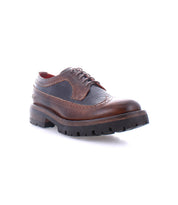 The Bed|Stü Lita K III is pictured against a white background, showcasing its brown leather brogue design with dark textured canvas and leather panels, red lining, decorative stitching, and a thick, rugged rubber sole.