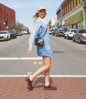 A woman in a blue dress, striped shirt, and tan hat crosses a city street lined with parked cars and brick buildings, her effortlessly chic look completed by vintage-inspired Bed|Stü Lita K III shoes that add a classic touch.