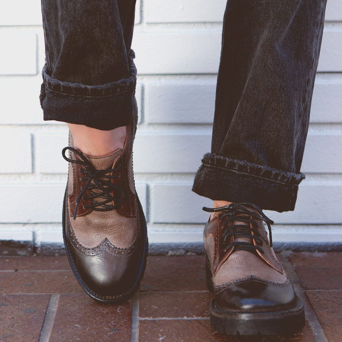 A person stands on a brick surface in front of a white brick wall, wearing black cuffed jeans and the Bed|Stü Lita K III brown leather vintage-inspired brogue shoes.
