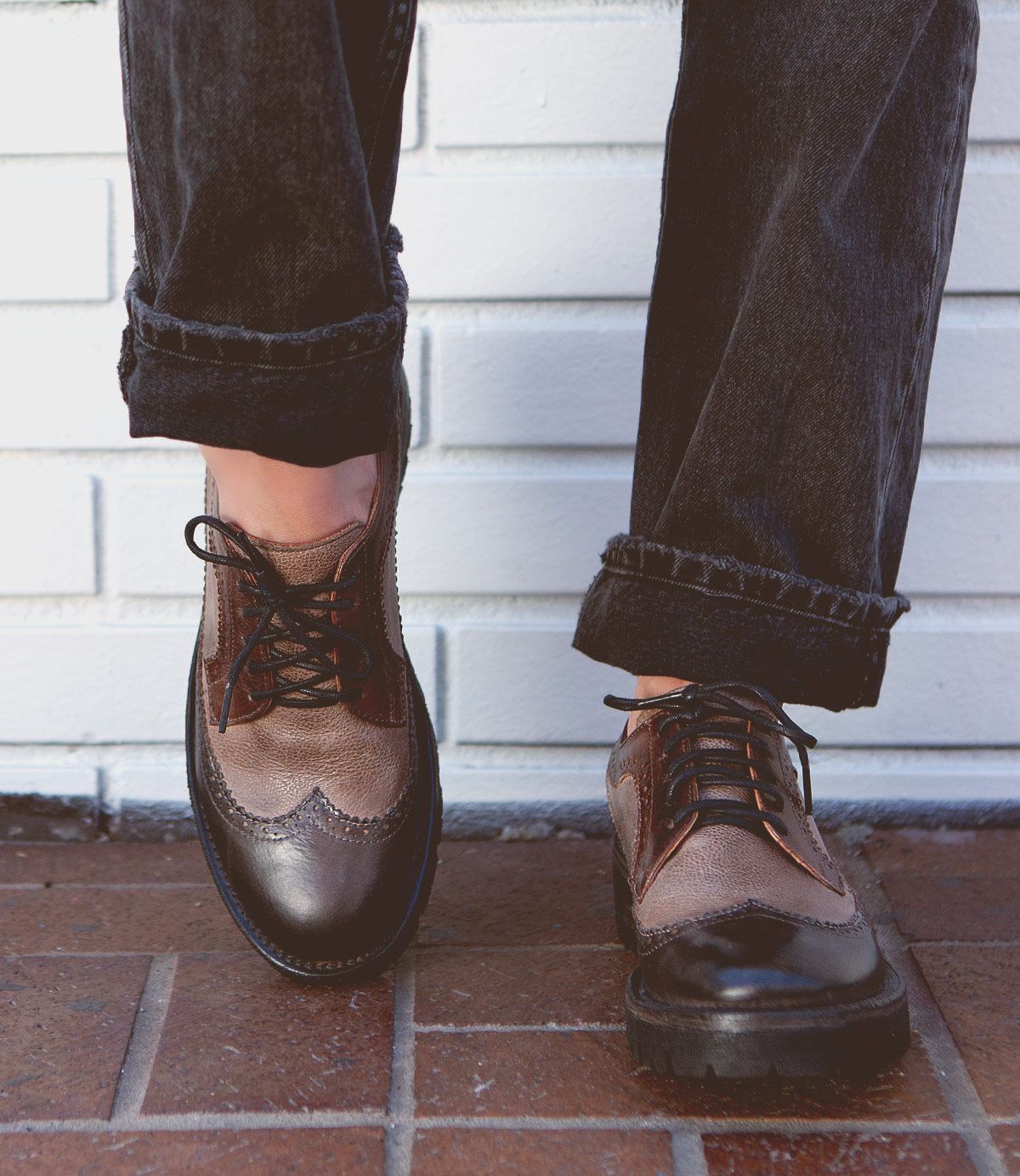A person stands on a brick surface in front of a white brick wall, wearing black cuffed jeans and the Bed|Stü Lita K III brown leather vintage-inspired brogue shoes.