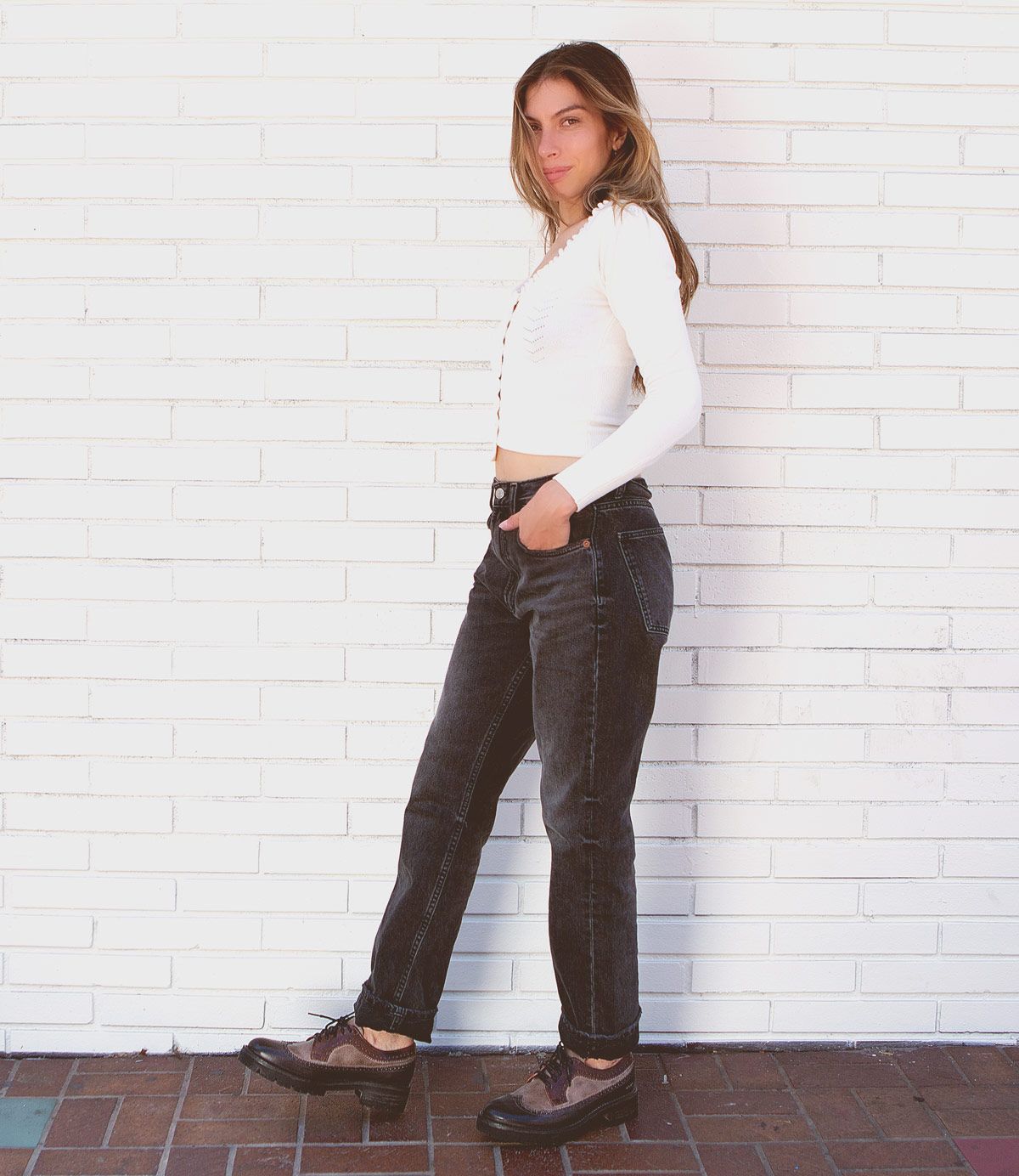 A person stands sideways against a white brick wall, wearing a white long-sleeve crop top, dark jeans, and the Bed|Stü Lita K III vintage-inspired platform shoes crafted from canvas and leather.