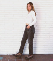 A person stands sideways against a white brick wall, wearing a white long-sleeve crop top, dark jeans, and the Bed|Stü Lita K III vintage-inspired platform shoes crafted from canvas and leather.