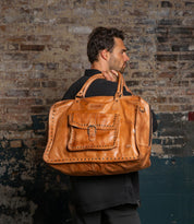A man in a black shirt holds the Bed|Stü Loaf— a large tan leather duffle with a front pocket— while standing against a textured brick wall.