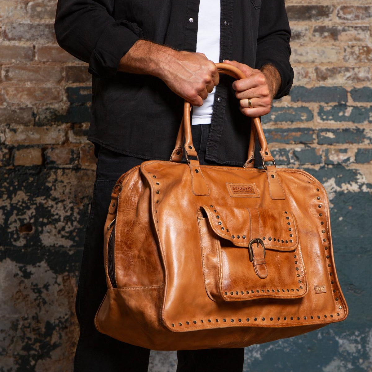 A person wearing a black shirt holds the Bed|Stü Loaf large tan leather duffle travel bag, featuring a front pocket and studded trim, while standing against a textured brick wall.