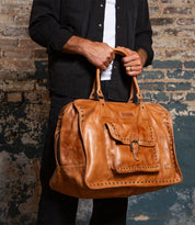 A person wearing a black shirt holds the Bed|Stü Loaf large tan leather duffle travel bag, featuring a front pocket and studded trim, while standing against a textured brick wall.