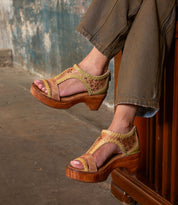 A person wearing wide-leg brown pants and the Bed|Stü Locket tan platform sandals—designed with cutout and braided details plus a cushioned footbed for extra comfort—sits indoors with legs crossed.