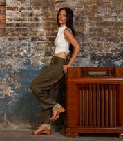 A woman in a white sleeveless top and olive pants leans against a vintage wooden radio before a weathered brick wall, her look completed by Bed|Stü's Locket sandals featuring laser-cut floral leather and a sculpted wooden heel.