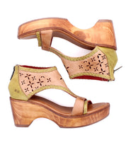 Two Bed|Stü Locket wooden platform sandals with tan laser-cut floral leather uppers, green accents, cushioned footbeds, open toes, cutout floral patterns, sculpted wooden heels, and back zippers are displayed side by side on a white background.