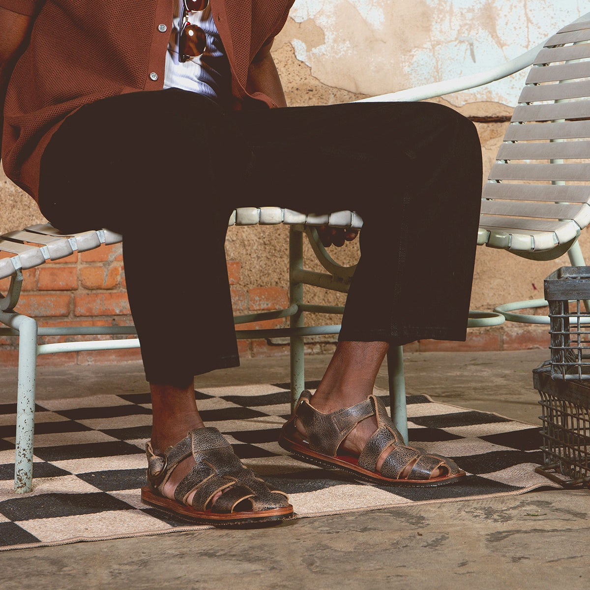 A person sits on a metal bench, dressed in a brown jacket and black pants, wearing Bed|Stü's Lone distressed leather sandals featuring an antique brass buckle and contoured footbed. Their legs are crossed on a checkered floor, with a textured wall in the background.