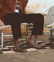 A person sits on a metal bench, dressed in a brown jacket and black pants, wearing Bed|Stü's Lone distressed leather sandals featuring an antique brass buckle and contoured footbed. Their legs are crossed on a checkered floor, with a textured wall in the background.