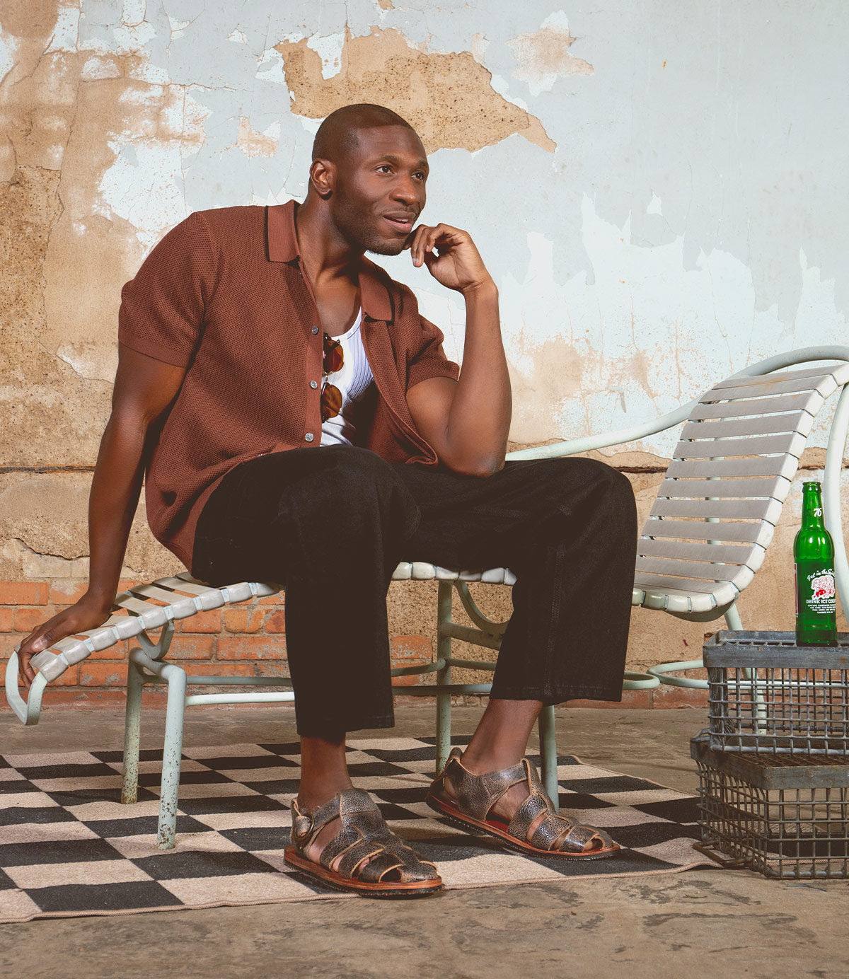A man in a brown shirt and black pants sits on a lounge chair, smiling, with a green bottle on a metal table beside him against a worn, textured wall. He wears the Bed|Stü Lone sandals—distressed leather with an antique brass buckle and contoured footbed—adding rugged character to the relaxed scene.