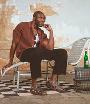 A man in a brown shirt and black pants sits on a lounge chair, smiling, with a green bottle on a metal table beside him against a worn, textured wall. He wears the Bed|Stü Lone sandals—distressed leather with an antique brass buckle and contoured footbed—adding rugged character to the relaxed scene.