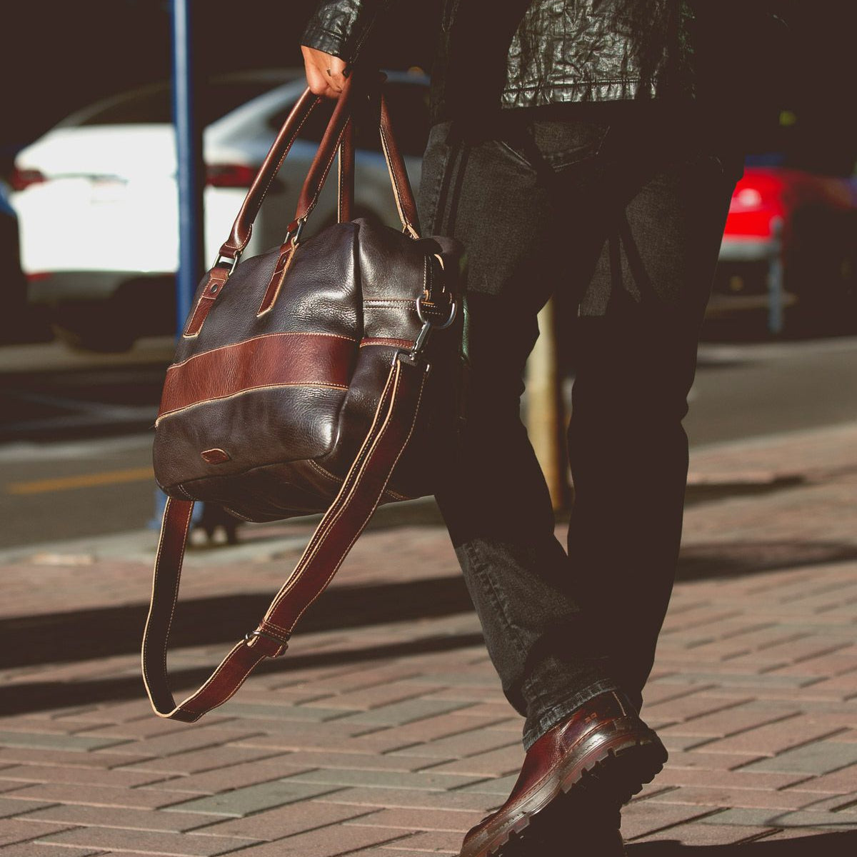 A person in dark clothes walks on a brick sidewalk, carrying the oversized Luke duffle by Bed|Stü, crafted from vegetable tanned leather and featuring a sturdy shoulder strap; parked cars are visible in the background.