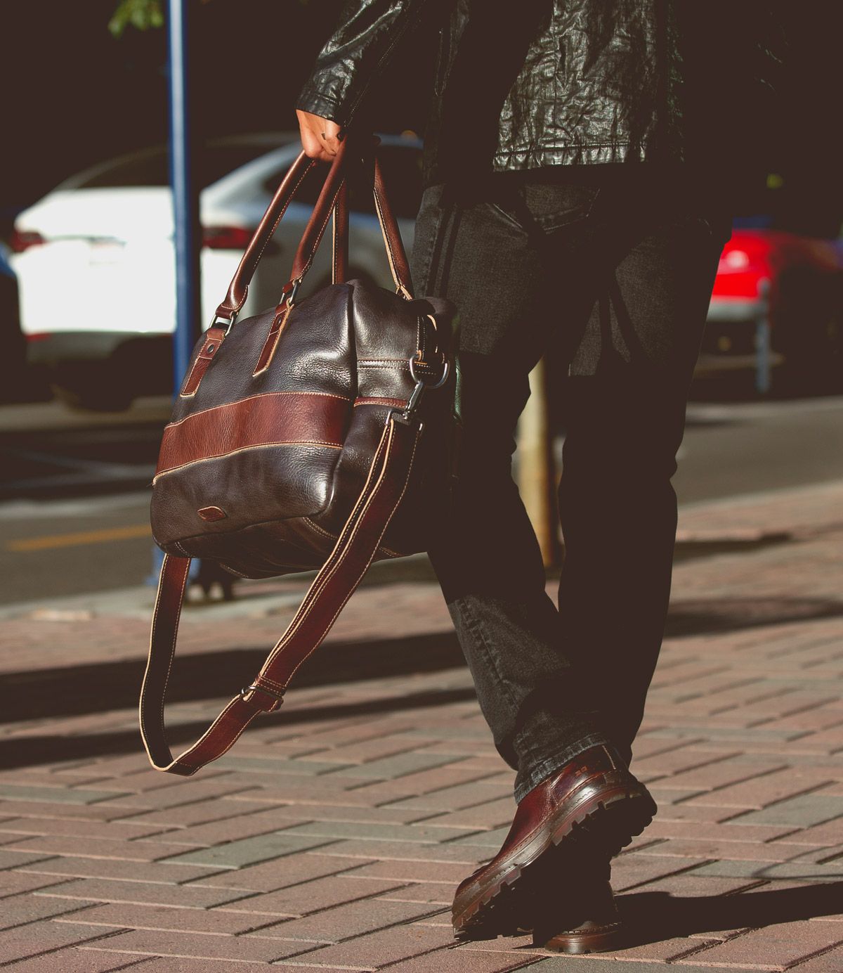 A person in dark clothes walks on a brick sidewalk, carrying the oversized Luke duffle by Bed|Stü, crafted from vegetable tanned leather and featuring a sturdy shoulder strap; parked cars are visible in the background.