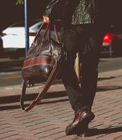 A person in dark clothes walks on a brick sidewalk, carrying the oversized Luke duffle by Bed|Stü, crafted from vegetable tanned leather and featuring a sturdy shoulder strap; parked cars are visible in the background.