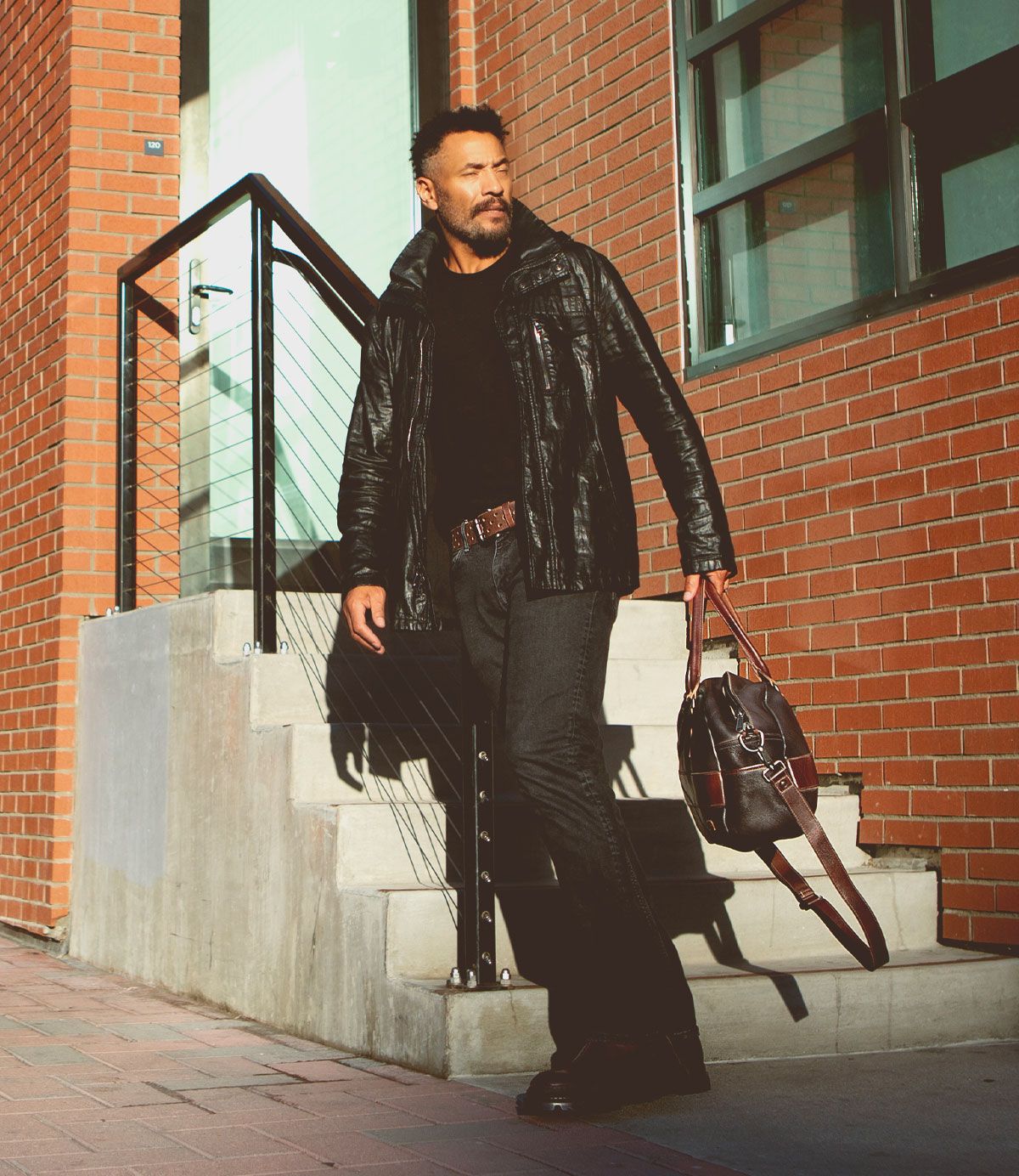 A man in a black jacket and dark jeans stands on steps near a brick building, holding the Bed|Stü Luke oversized duffle crafted from vegetable tanned leather and looking to the side in sunlight.