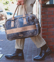A person walks on a sidewalk wearing tan pants and black boots, carrying the Bed|Stü Luke oversized duffle made from vegetable tanned leather, featuring tan straps and a beige stripe.