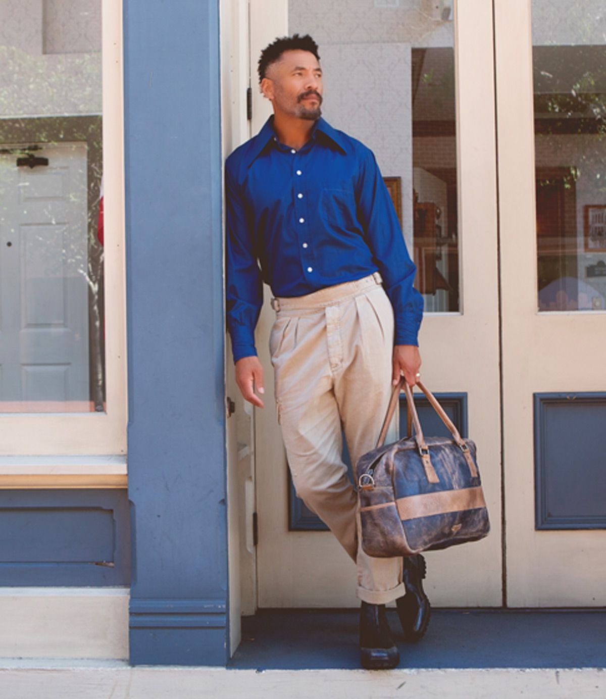 A man in a blue shirt and beige pants stands outside a building, leaning against the door frame while holding the Luke oversized duffle by Bed|Stü, crafted from vegetable tanned leather.