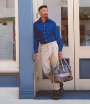 A man in a blue shirt and beige pants stands outside a building, leaning against the door frame while holding the Luke oversized duffle by Bed|Stü, crafted from vegetable tanned leather.