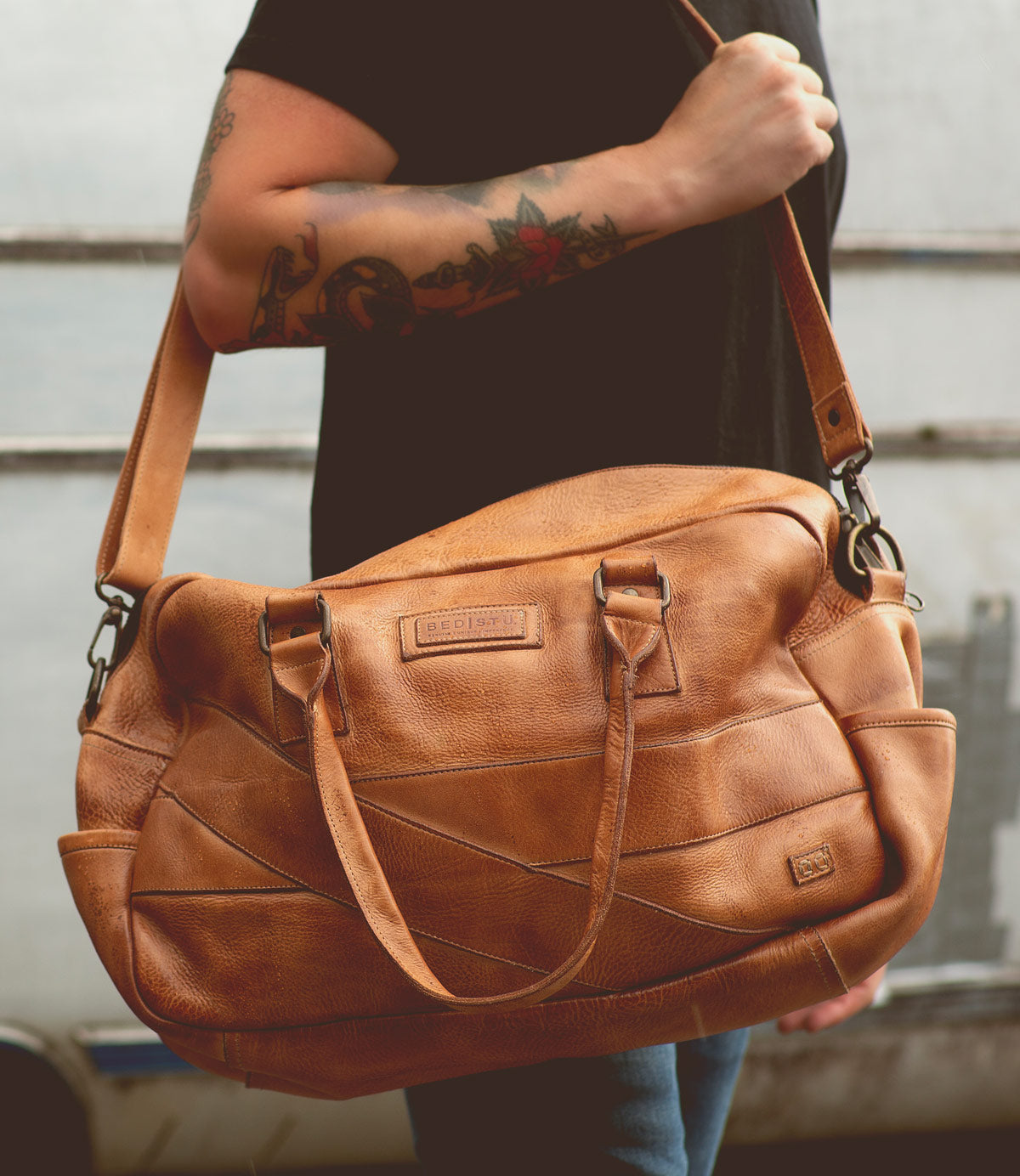 A person with tattooed arms holds the Bed|Stü Luke, an oversized duffle crafted from vegetable tanned leather, by the shoulder strap—showcasing a stylish choice in travel bag.