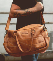 A person with tattooed arms holds the Bed|Stü Luke, an oversized duffle crafted from vegetable tanned leather, by the shoulder strap—showcasing a stylish choice in travel bag.